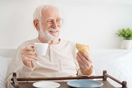 Photo of aged man happy positive smile dream look empty space have breakfast on bed drink coffee eat croissant homeの写真素材