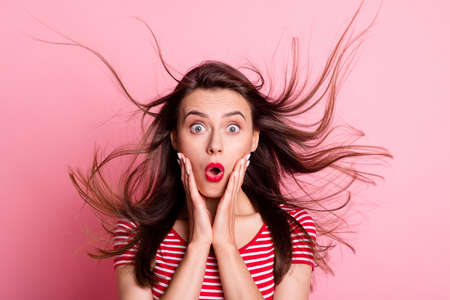 Portrait of young attractive shocked amazed surprised girl hold hands cheeks with flying hair isolated on pink color backgroundの写真素材