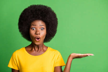 Photo of amazed brunette wavy haired dark skin woman hold hand empty space isolated on green color backgroundの写真素材