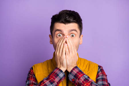 Photo of frightened impressed young man wear yellow sleeveless jacket arms cover face isolated violet color backgroundの写真素材