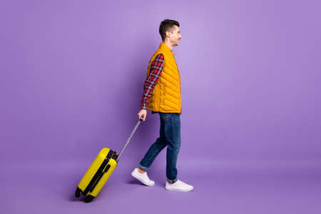 Full length photo of cheerful young man wear yellow sleeveless jacket holding suitcase walking...