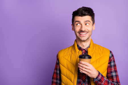 Photo of cute cheerful young guy dressed yellow vest holding brown cup looking empty space isolated purple color backgroundの写真素材