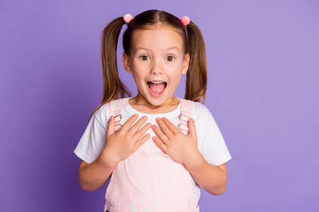 Photo of amazed cute little girl gesture show ask me wear pink overall dress isolated on violet color backgroundの写真素材