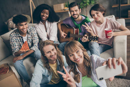 Photo portrait of funny students sitting on sofa in hostel taking selfie with smartphone eating pizza playing guitarの写真素材