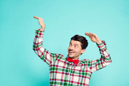 Photo of funky guy raise hands look side empty space shiny smile wear checkered shirt isolated blue color backgroundの写真素材