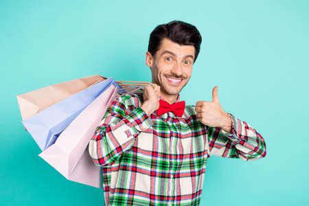Photo of customer guy hold store bags raise thumb up wear checkered shirt bow isolated blue color backgroundの写真素材
