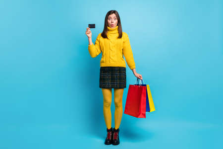 Full length photo of cute shocked young lady wear yellow sweater holding credit card bargains isolated blue color backgroundの写真素材