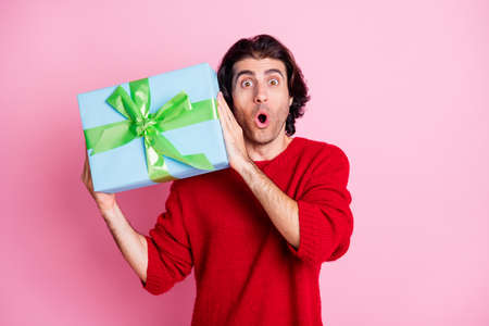 Photo portrait of shocked man shaking present near ear guessing whats inside isolated on pastel pink colored backgroundの写真素材