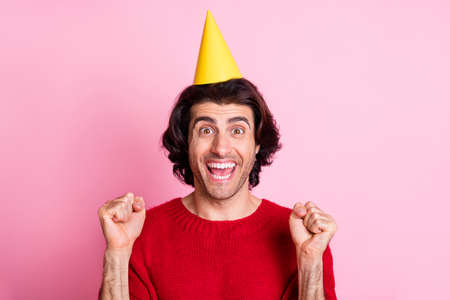 Photo portrait of celebrating man with two raised fists wearing yellow party cone isolated on pastel pink colored backgroundの写真素材