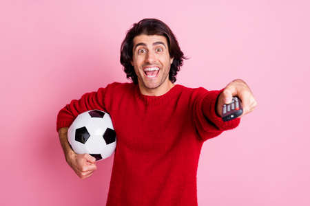Photo portrait of man with football changing tv channels with remote control isolated on pastel pink colored backgroundの写真素材