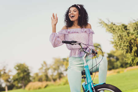 Portrait of excited positive dark skin lady sit on bike look interested far arm palm wave good mood sightseeing outdoorsの写真素材