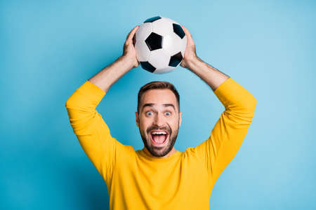 Photo of young crazy happy positive excited cheerful man hold football ball above head isolated on blue color backgroundの写真素材