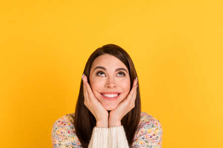 Photo of cheerful positive young woman hold hands cheeks look up empty space isolated on yellow color backgroundの写真素材