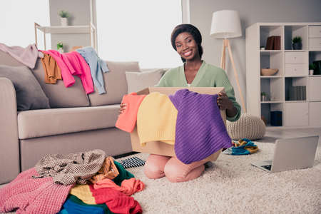 Portrait of attractive cheerful girl sitting on floor folding clothes gathering in box economy...