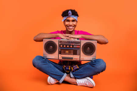 Full length body size photo of young man with sitting keeping boom box smiling isolated vivid orange color backgroundの写真素材