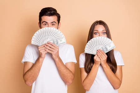 Photo of charming cute excited husband wife wear white t-shirts dollars cover face isolated beige color backgroundの写真素材