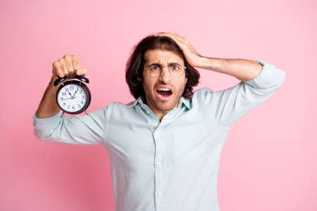 Photo of stressed funny guy hold clock oversleep wear spectacles blue shirt isolated on pink color...
