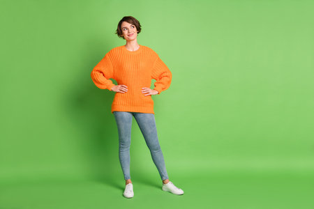 Full length photo of curious happy young lady look empty space hold hands waist isolated on green color backgroundの写真素材