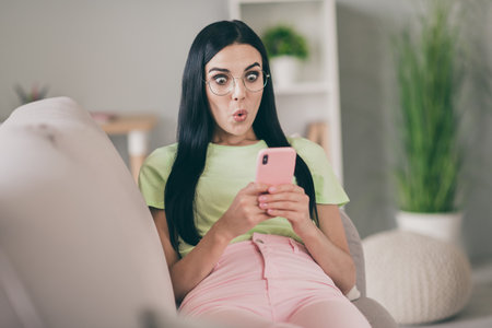 Photo of funny shocked young lady lay couch hold cellphone surprised face open mouth wear specs...