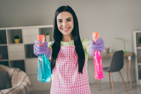 Portrait of pretty cheerful long-haired maid holding in hands using bottles pulverizers cleanup at light interior living-roomの写真素材