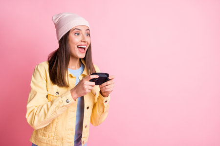 Portrait of beautiful young lady playing games gamepad in hands open mouth wear cap isolated on pink...