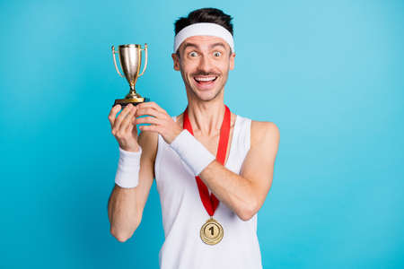 Photo of young cheerful sportive man happy positive smile win competition hold trophy isolated over blue color backgroundの写真素材