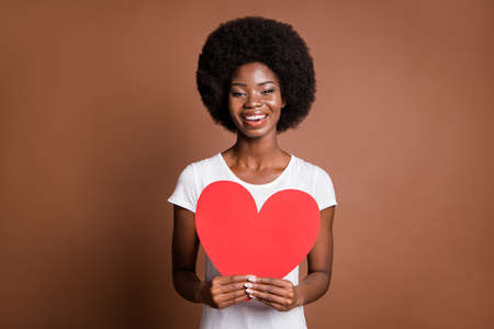 Photo of cheerful romantic girl hold red paper heart toothy smile wear white t-shirt isolated brown color backgroundの写真素材