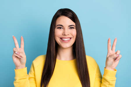 Photo of positive charming lady show two v-signs beaming smile wear yellow jumper isolated blue...