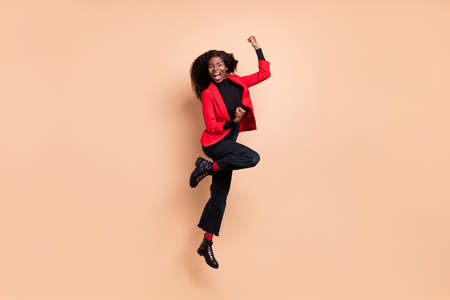 Photo of excited lucky curly dark skin woman dressed red blazer jumping rising fists isolated beige color backgroundの写真素材