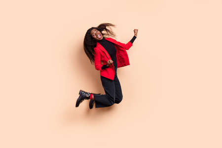 Photo of lucky funny curly dark skin woman dressed red blazer jumping rising fists isolated beige...