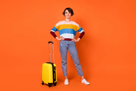 Photo of charming cute young lady dressed striped pullover luggage standing arms waist isolated orange color backgroundの写真素材