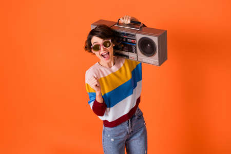 Photo of funky lucky young woman wear striped sweater eyewear rising fist holding boombox isolated orange color backgroundの写真素材