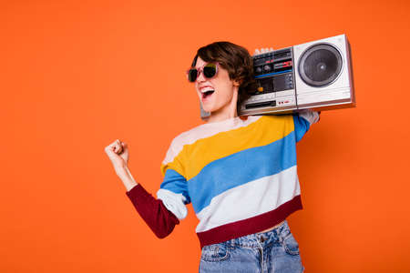 Photo of delighted funny girl fist celebrate look empty space hold boom box isolated on orange color backgroundの写真素材