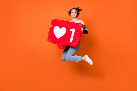 Photo of cute sweet young woman wear striped sweater jumping holding red like sign isolated orange color backgroundの写真素材