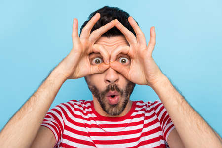 Photo of funny shocked young guy wear red showing spectacles fingers cover eyes isolated blue color...
