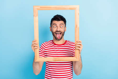 Photo of childish funny young guy wear red outfit holding looking wooden frame showing tongue isolated blue color backgroundの写真素材