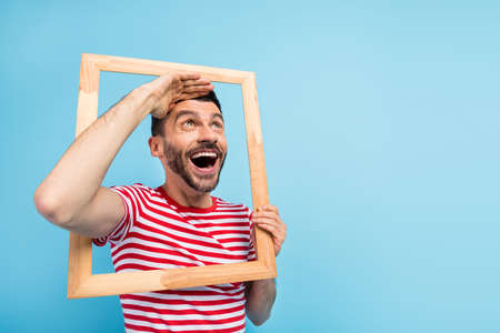 Photo of impressed cute young man dressed striped t-shirt looking brown frame arm forehead empty space isolated blue color backgroundの写真素材