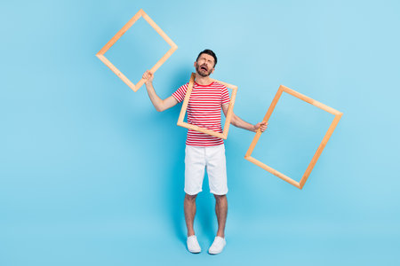 Photo of unhappy upset young guy wear red outfit holding three wooden frames crying isolated blue...