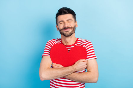 Photo of cute dreamy young man dressed striped t-shirt embracing red paper heart closed eyes isolated blue color backgroundの写真素材