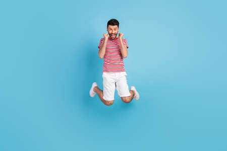 Full length photo of funky lucky young man dressed striped t-shirt jumping high rising fists isolated blue color backgroundの写真素材