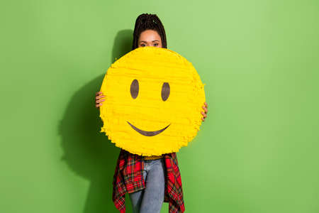 Portrait of nice girl holding in hands closing face large yellow cute childish smiley isolated over bright green color backgroundの写真素材