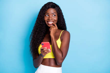 Photo of dark skin girl dressed yellow singlet holding modern device biting finger looking empty...