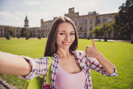 Photo of charming happy young woman make thumb up selfie camera wear plaid shirt outside in campus outdoorsの写真素材