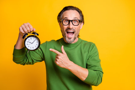 Photo of excited man hold alarm clock open mouth direct finger wear eyewear green sweater isolated yellow color backgroundの写真素材