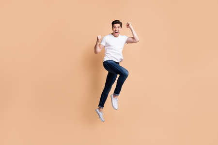 Full size profile photo of hooray brunet man jump wear t-shirt jeans sneakers isolated on beige backgroundの写真素材