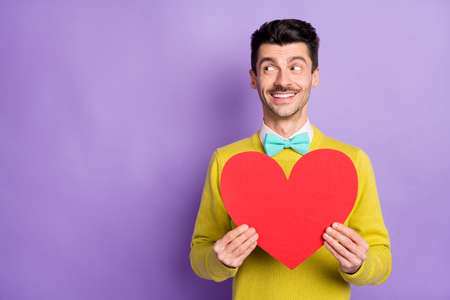 Portrait of brunet optimistic guy hold paper heart look empty space wear yellow sweater isolated on...