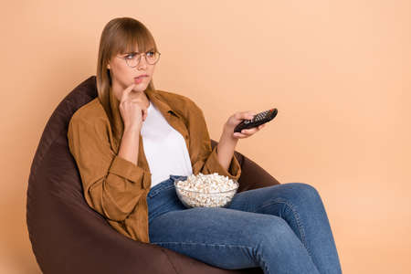 Photo of puzzled charming young lady bite lip switch channel sit chair eat popcorn isolated on pastel beige color backgroundの写真素材