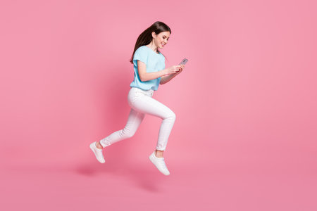 Full length profile side photo of girl jump use smartphone run wear t-shirt trousers isolated pastel color backgroundの写真素材