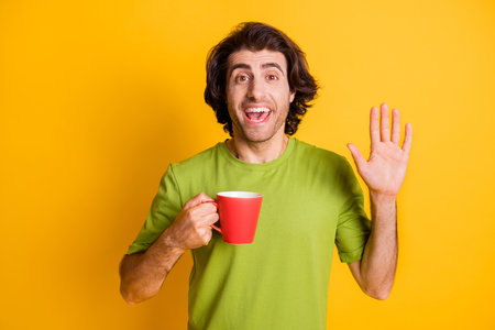 Portrait photo of man keeping cup of coffee waving with hand greeting isolated on vibrant yellow color backgroundの写真素材