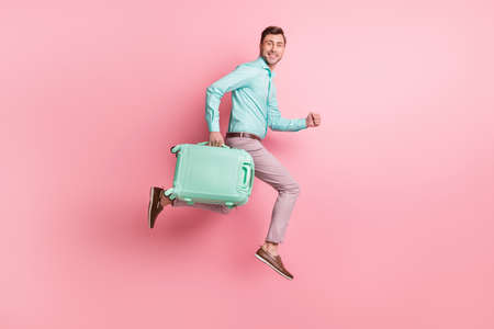 Full size profile side photo of brunette man hold luggage trip run jump wear teal shirt pink pants isolated on pastel color backgroundの写真素材